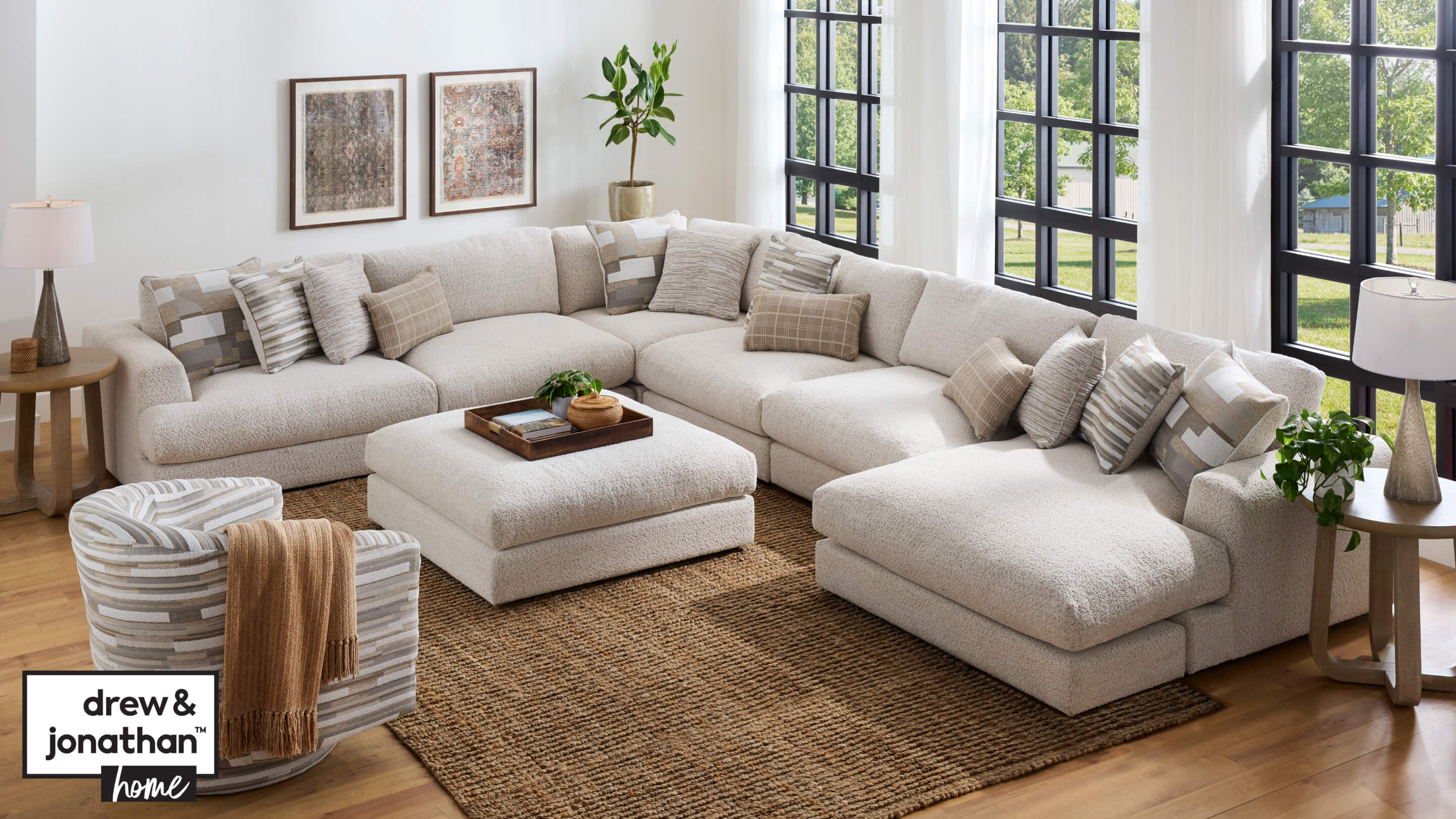 Living room scene with Montecito sectional in off white color with ottoman. Dark brown wood tray with green plant and book on top of ottoman. Accent chair in multi colored gray and tan pattern to the left of the sectional. Tan jute area rug beneath the sectional and ottoman. Shaw end tables on either end of the sectional with neutral colored lamps.