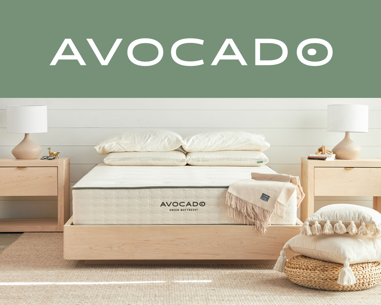 Avocado logo at the top, below that is a bedroom set up, with a light wood bed frame and avocado mattress. Either side of the bed features matching light wood nightstands and cream lamps. A stack of accent pillows and a braided pouf lay on the floor to the right of the bed.