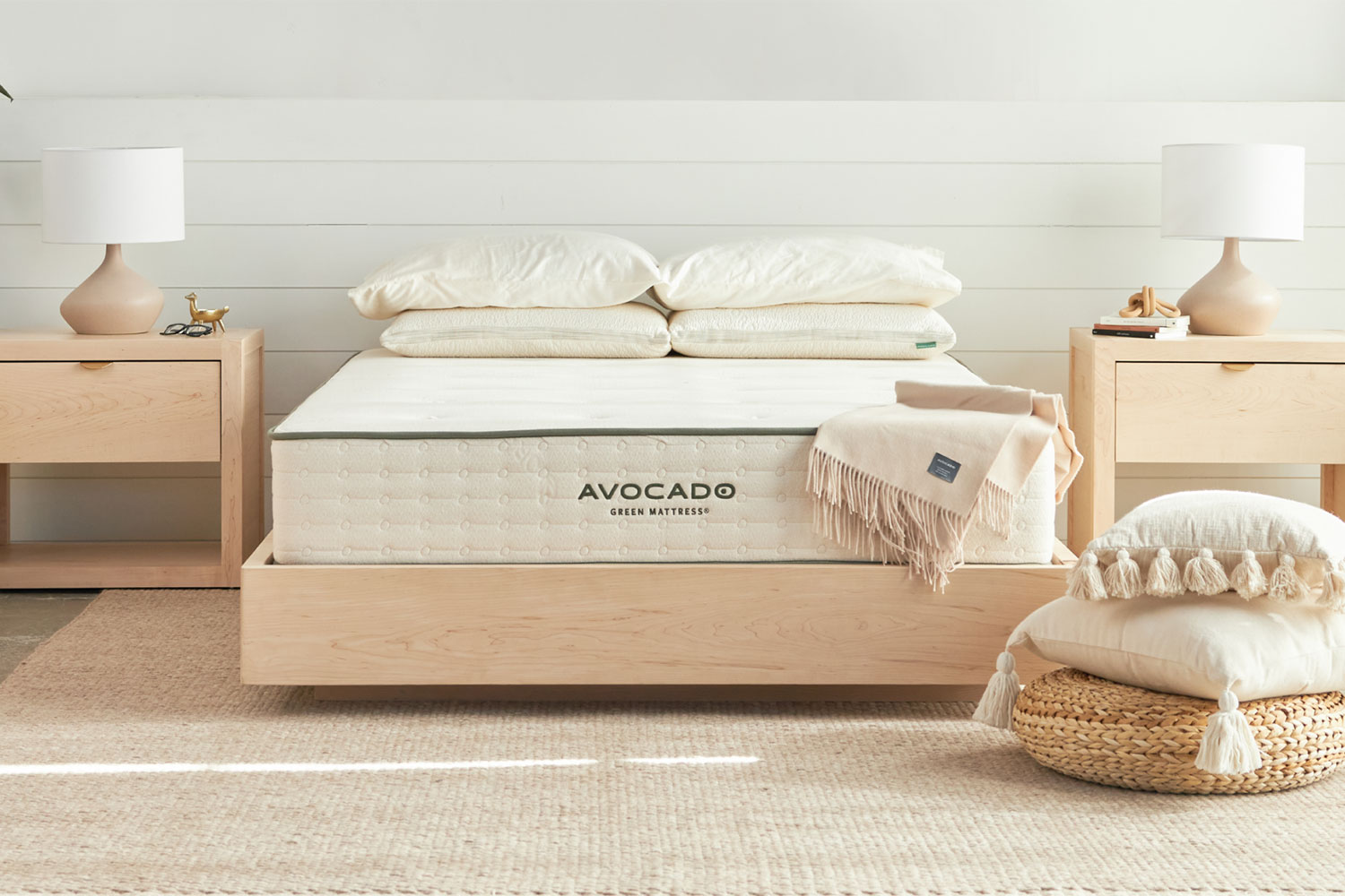Bedroom setting with Avocado green mattress, on a light wood bed frame, with matching nightstands on either side of the bed. Cream bedside lamps on each nightstand, and a stack of accent pillows on the ground to the right of the foot of the bed.